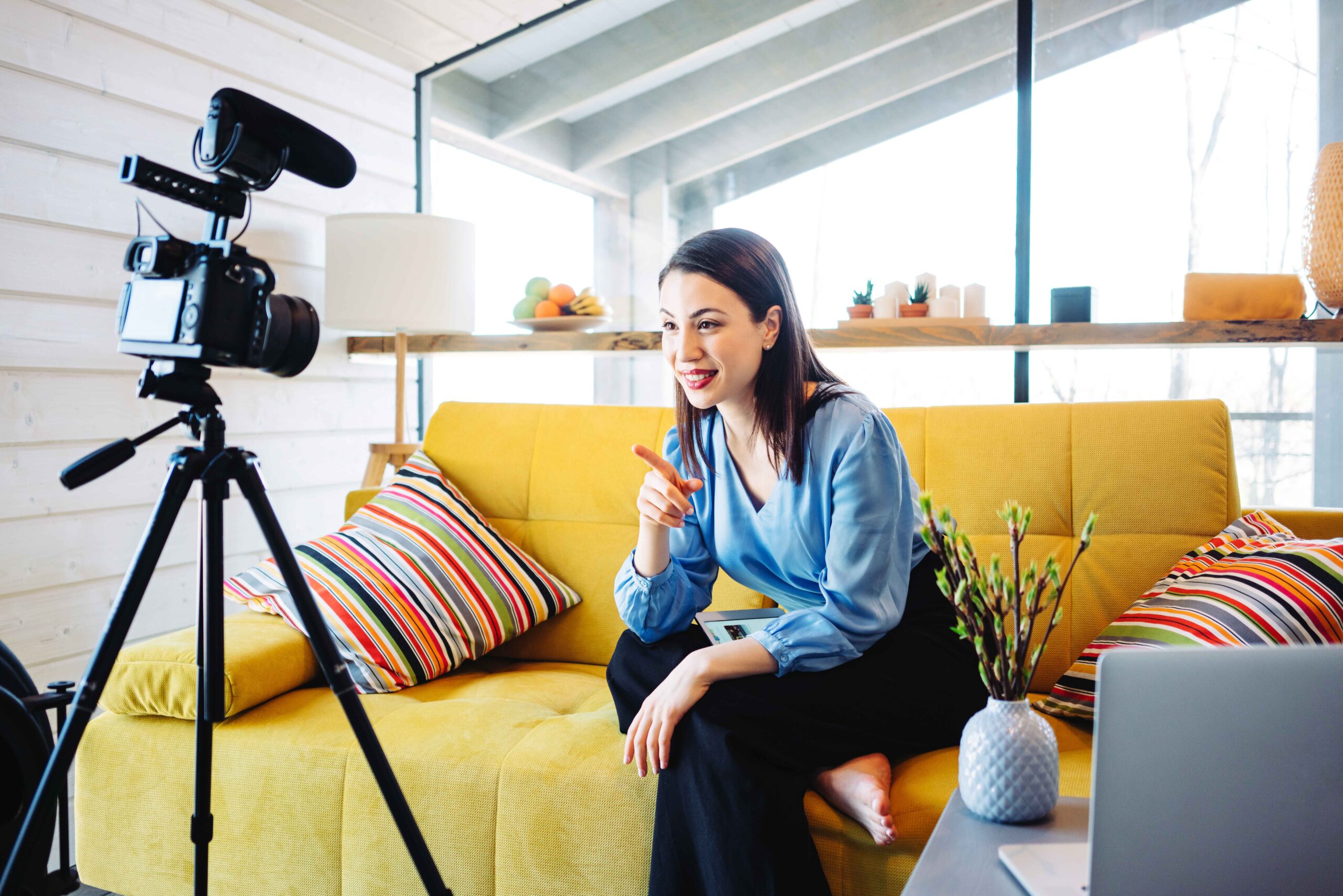 Woman seated on a yellow couch taping video content with a remote setup.