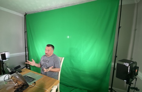 Man at a desk in front of a green screen, recording a presentation on a laptop.